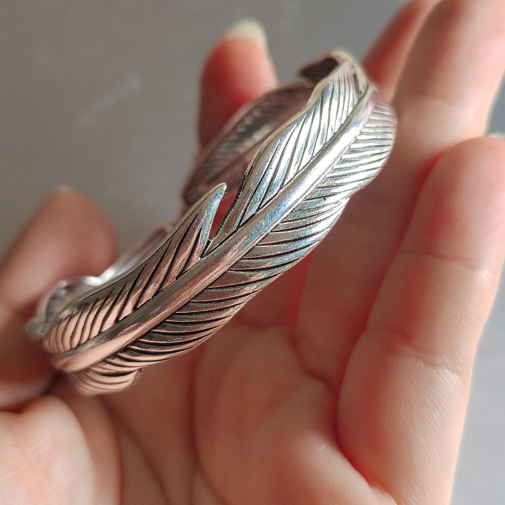 Silver feather-shaped bracelet held in a hand against a neutral background