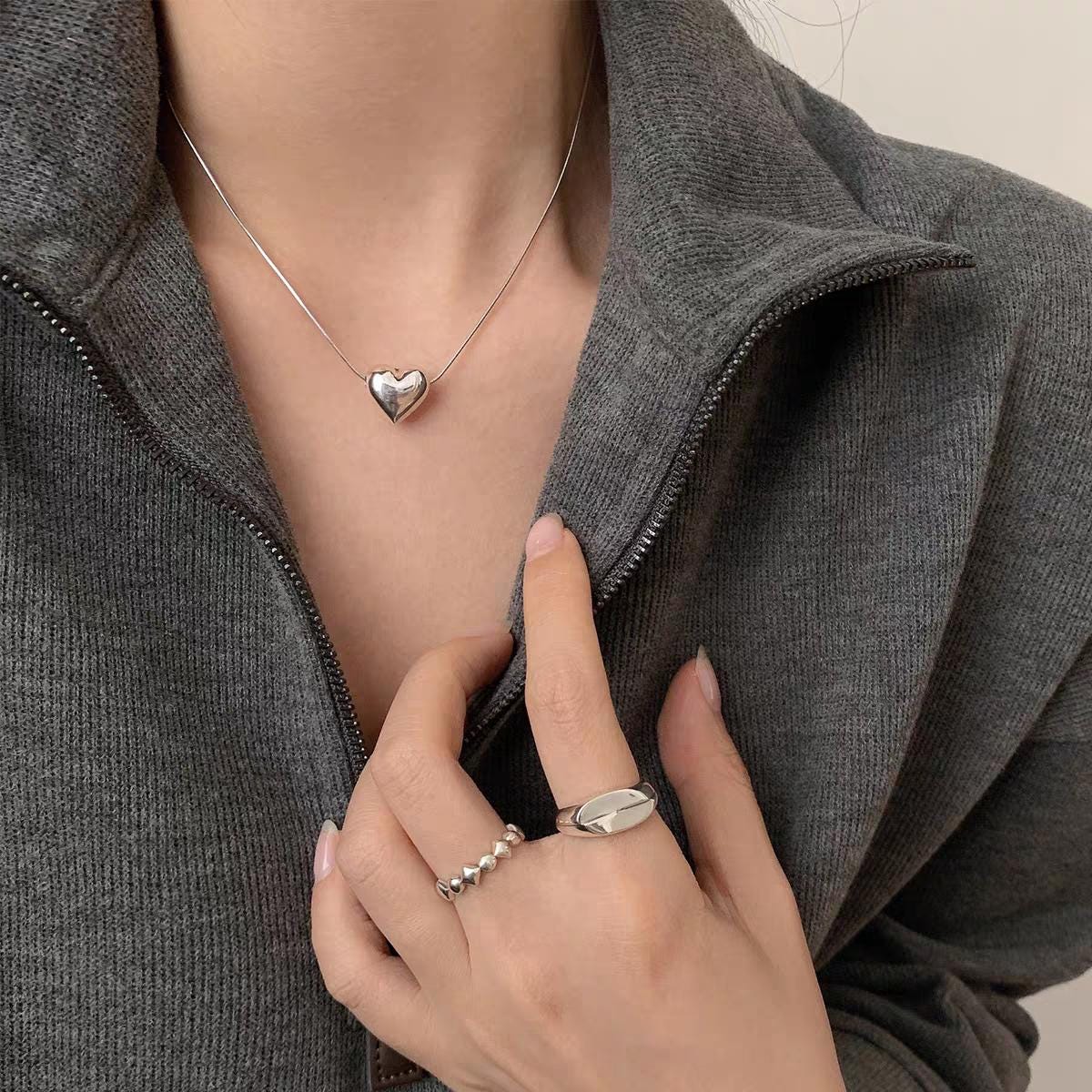 Person wearing a silver heart-shaped necklace and multiple rings on a neutral background
