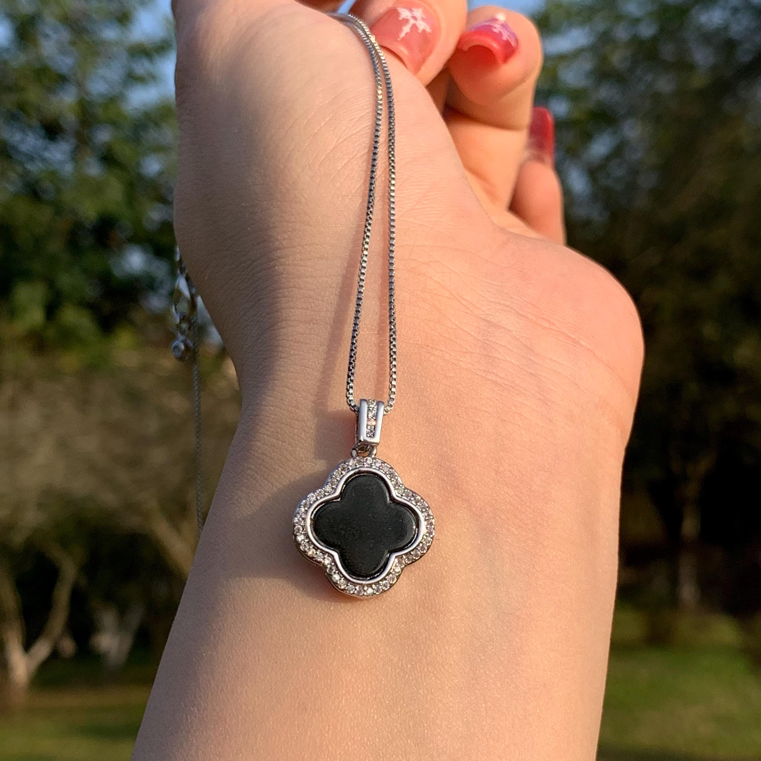 Hand holding a necklace with a black floralclover pendant against a blurred natural background