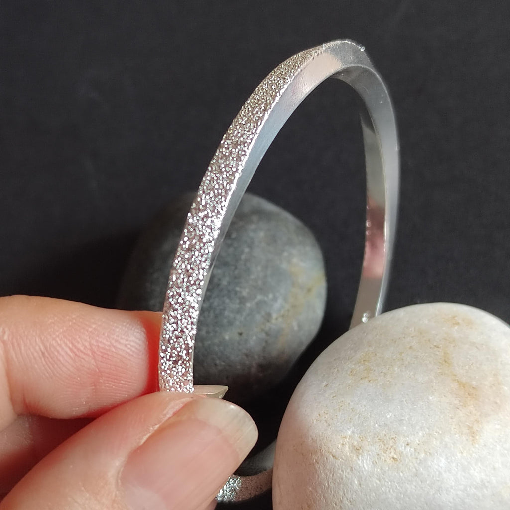 Silver bracelet held by a hand with pebbles in the background