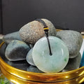 Green jade pendant on a black cord with pebbles on a reflective surface