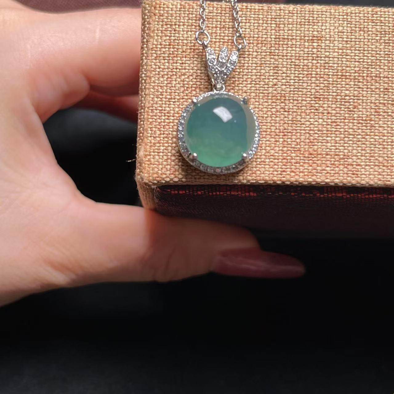 Green gemstone pendant on a chain held by a hand against a textured brown background