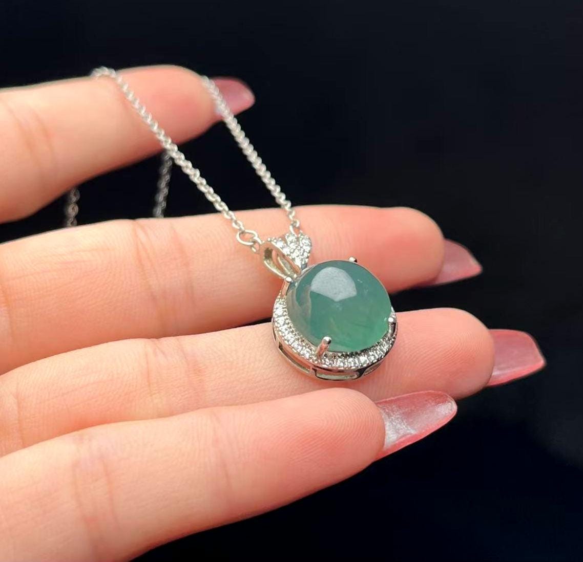 Green gemstone pendant on a silver chain held by a hand against a black background