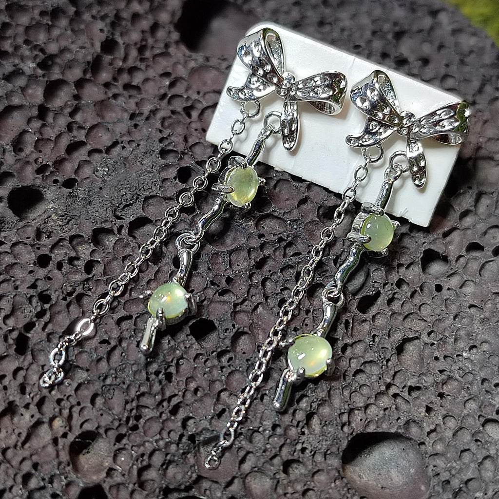 Silver earrings with bow designs and green gemstones on a textured surface