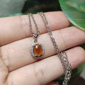 Silver necklace with a pendant featuring an orange stone held between fingers against a blurred green leaf background