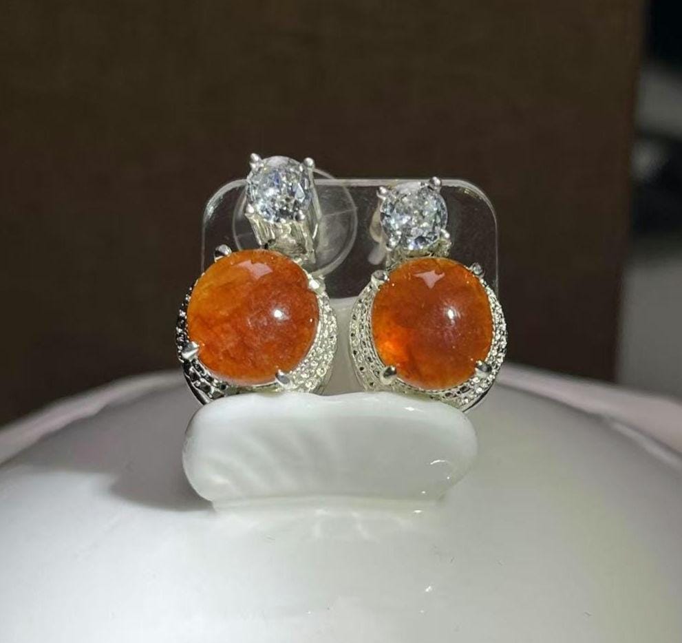 Pair of earrings with orange gemstones and silver settings on a white stand.