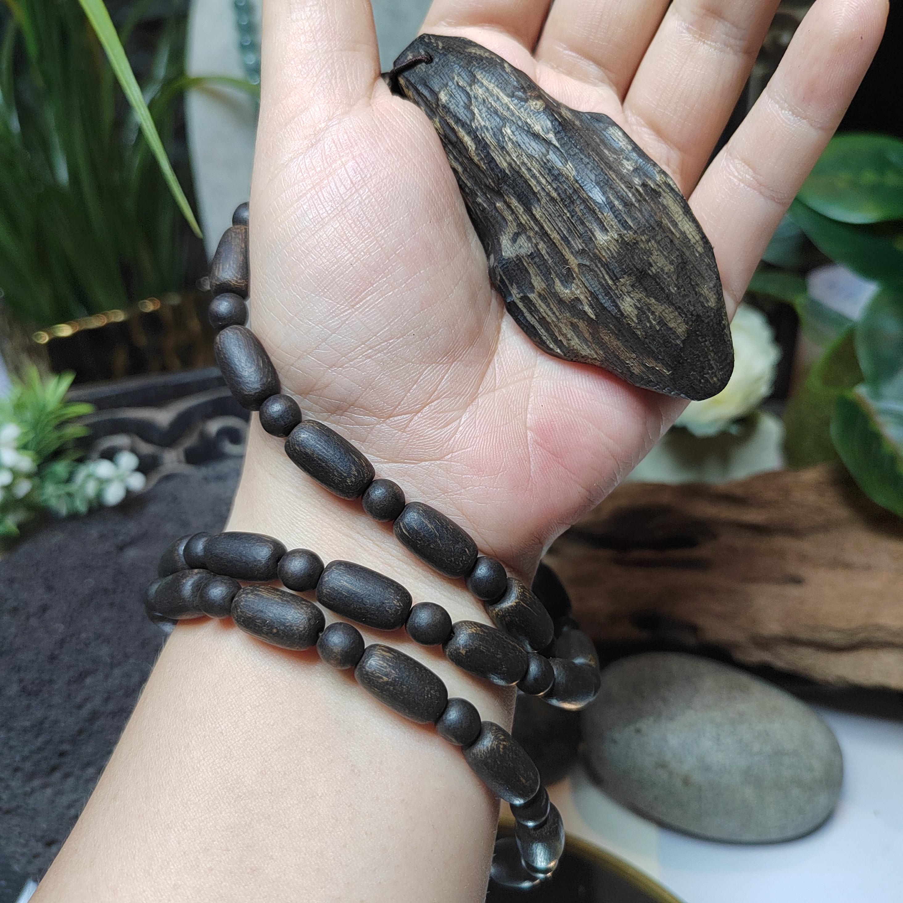Hand wearing a black beaded bracelet with a wooden pendant, surrounded by plants.