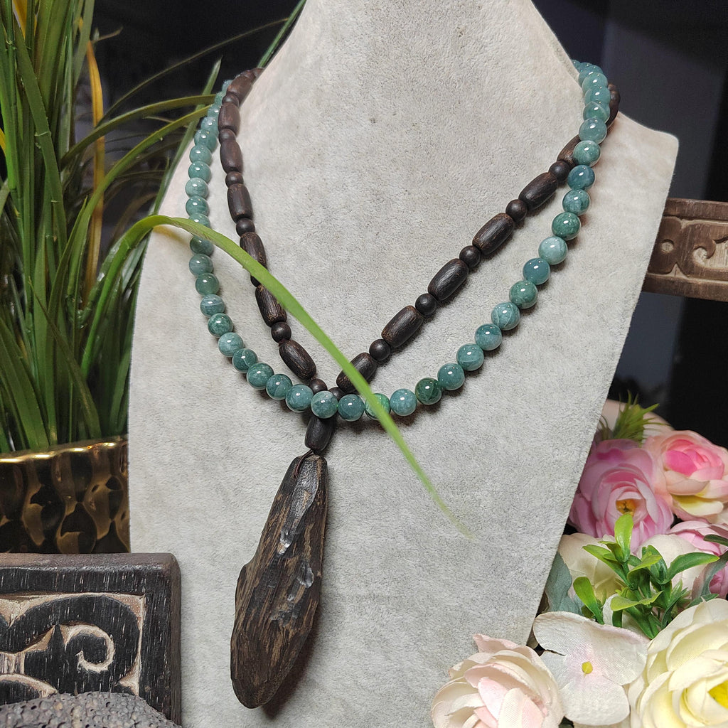 Necklace with green beads and a wooden pendant on a gray stand with flowers and plants in the background