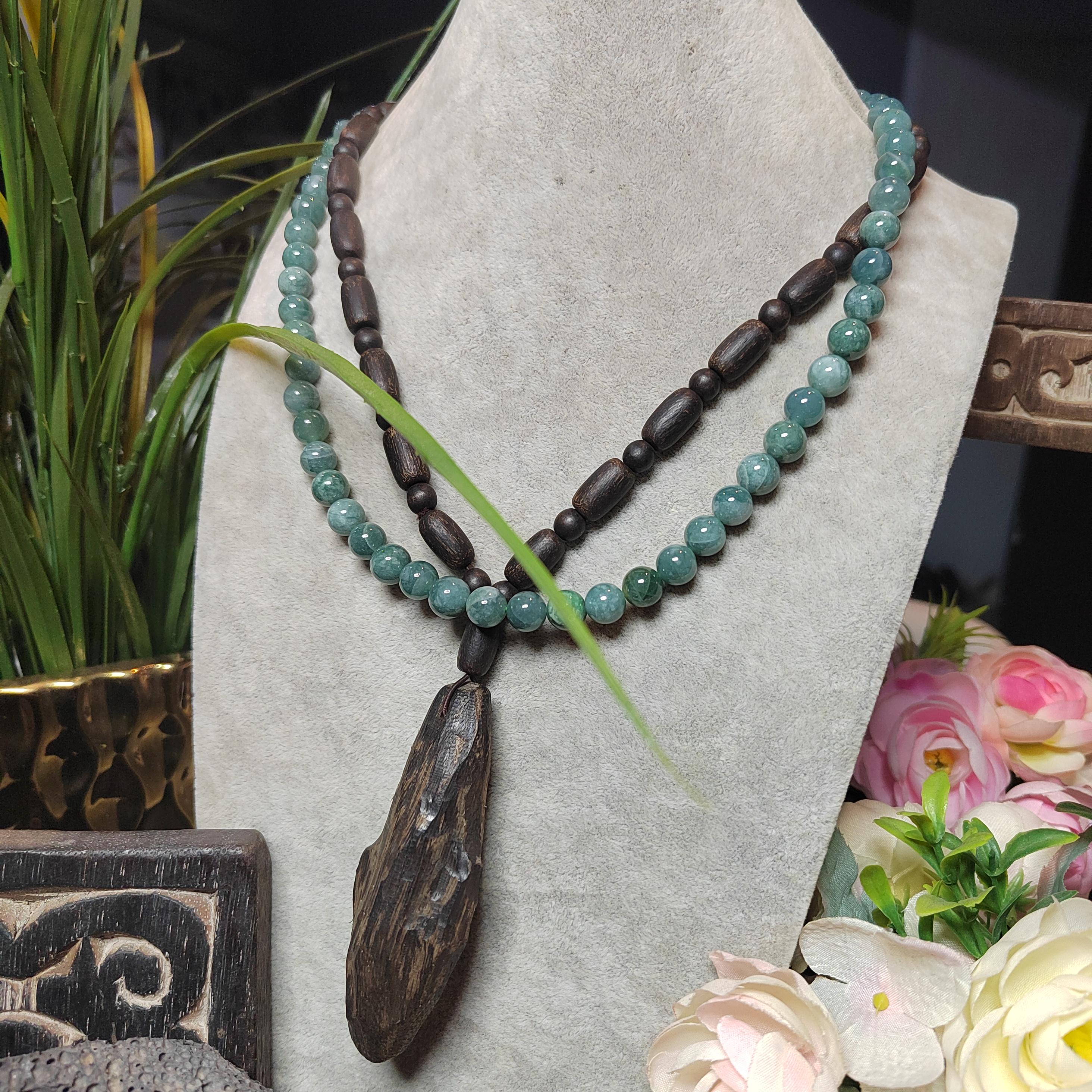 Necklace with green beads and a wooden pendant on a gray stand with flowers and plants in the background