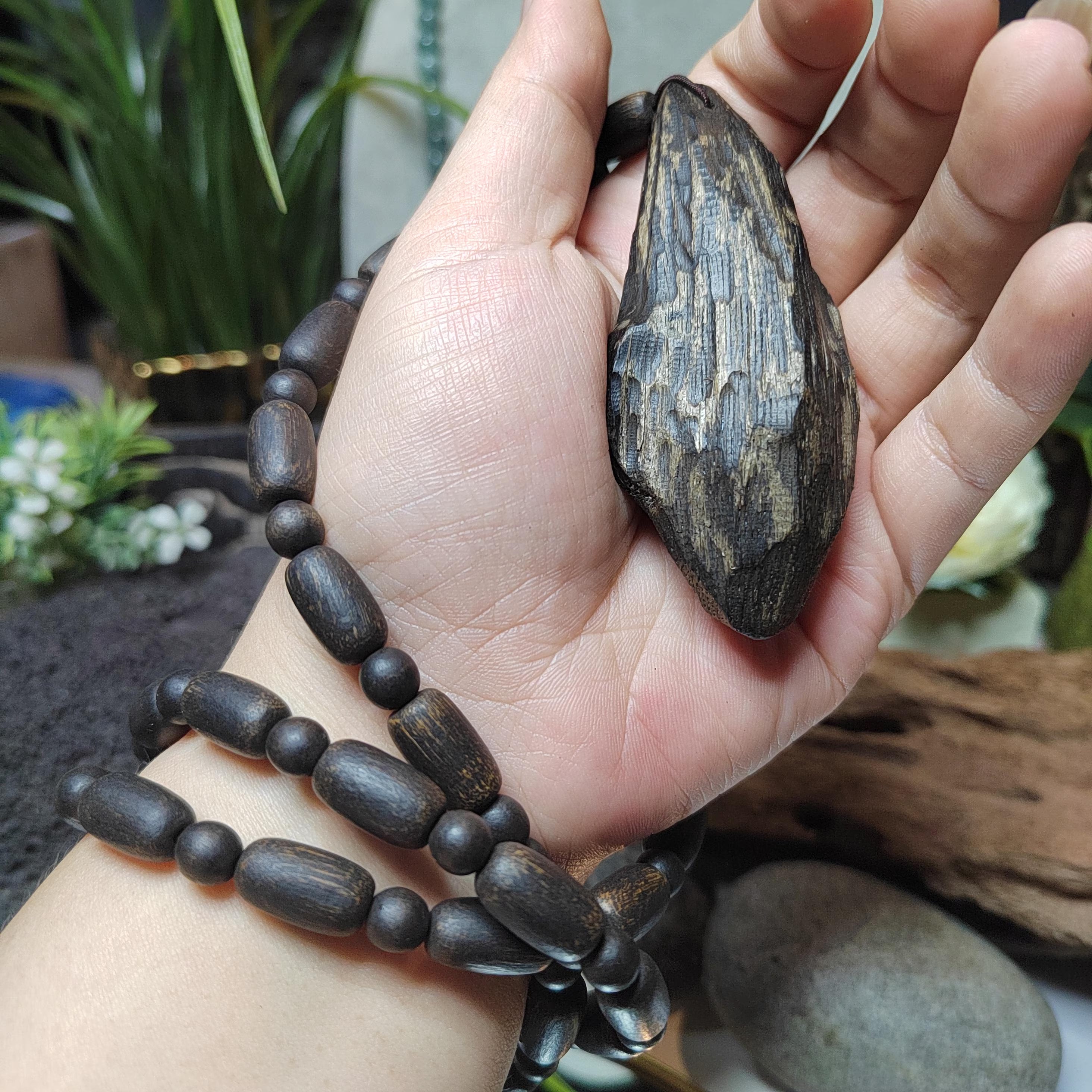 Hand holding a dark brown stone and a black beaded necklace with a blurred natural background