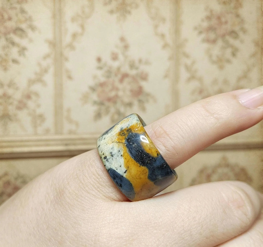 Ring with a multicolored stone on a finger against a floral-patterned wall.