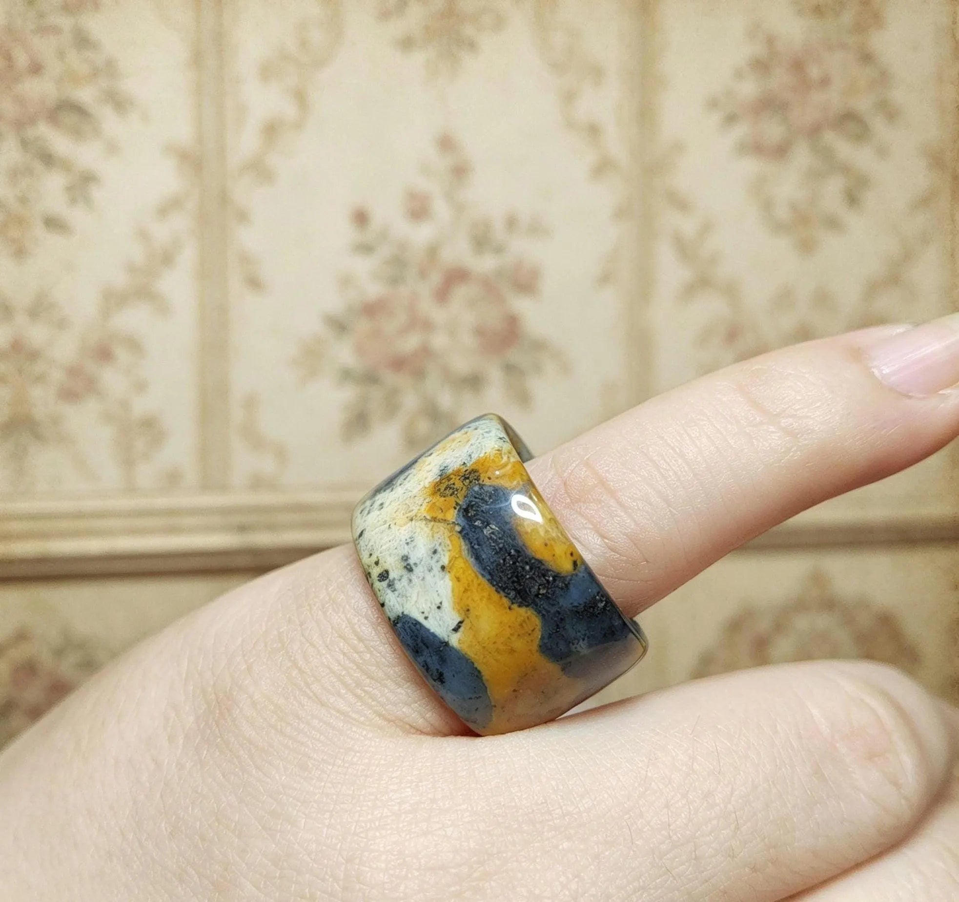 Ring with a multicolored stone on a finger against a floral-patterned wall.